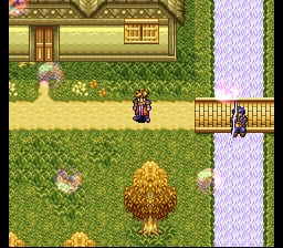 A top-down perspective shows a character sprite in the center of a grassy outdoor area with a stone path dividing the screen vertically. Yellow flowers and rocks are scattered across the grass. A building with a brown roof appears in the upper left, and a wooden fence structure runs along the right edge. The pixel art uses a 16-bit color palette with green grass, lavender-colored stone tiles, and detailed foliage in the background, typical of SNES-era RPG graphics.