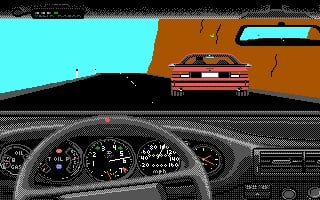 A first-person driving view from inside a car cockpit showing a detailed instrument dashboard with speedometer, tachometer, and other gauges across the lower third of the screen. A red sports car is visible on a desert highway ahead, approaching from the distance. The road curves through a brown/tan landscape under a cyan sky, with a black road surface and brown cliff walls flanking both sides. The graphics display pixelated 16-bit style rendering typical of late 1980s DOS racing games.