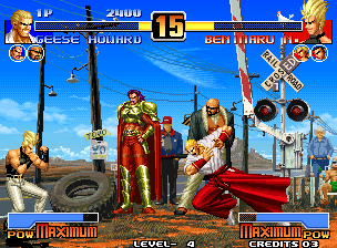 A fighting game match screen displays two characters in combat stance on a junkyard stage with stacked tires and industrial structures in the background. The top UI shows player names, health bars, and a round counter displaying "15". Character portraits appear in the upper corners, and a "POWER MAXIMUM" indicator is visible at the bottom left. The arcade sprite graphics use pixel art with a bright daytime setting and blue sky parallax background. Fighter sprites are rendered in mid-frame animation poses typical of 1990s 2D fighting games.