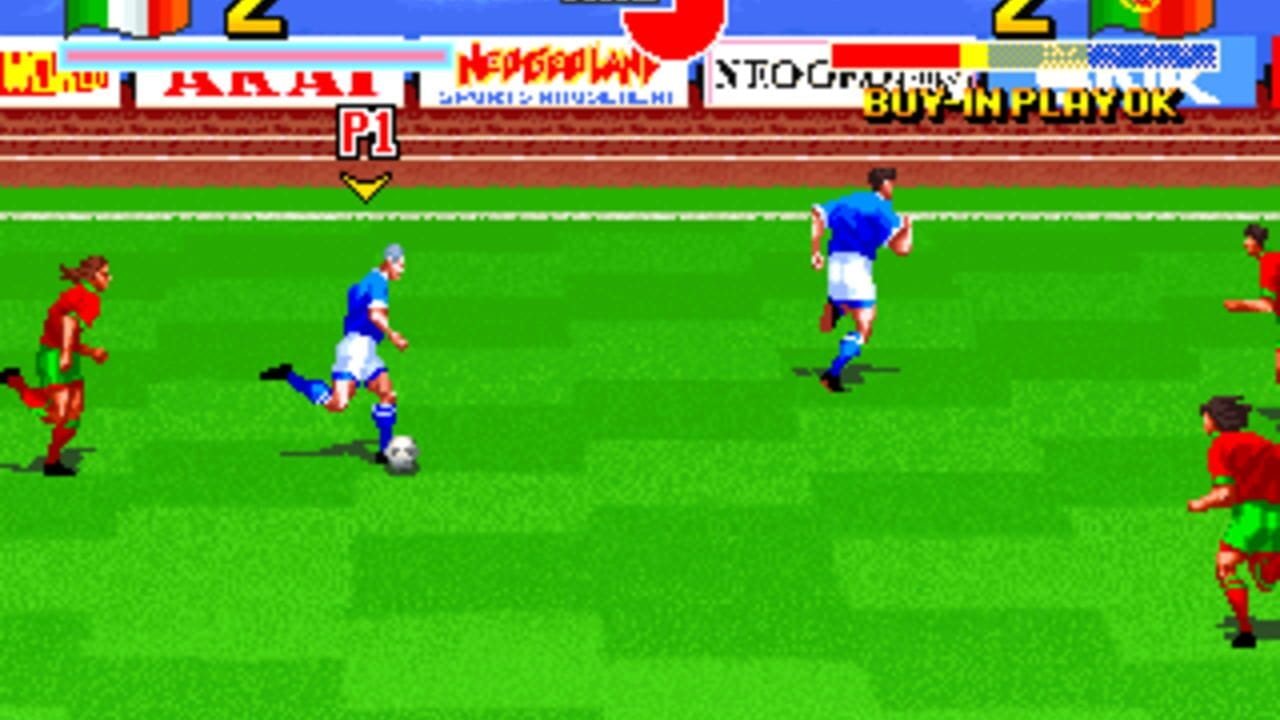 A soccer match in progress on a bright green field with players in blue and white uniforms controlling the ball against red-uniformed opponents. The top of the screen displays a brown stadium barrier, country flags, score indicators, and text reading "P1" with a yellow downward arrow. Multiple red players are positioned around the field edges while blue team members maneuver near the center, with a white soccer ball visible near a blue player's feet. The arcade interface shows simplified pixel-art character sprites typical of mid-1990s arcade graphics.