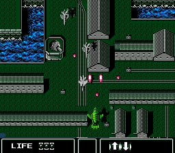 A top-down isometric view shows a green-toned street intersection with white building outlines and gridded pavement. A red and white vehicle occupies the center of the road. Blue water or liquid appears in the upper-left corner. The HUD displays "LIFE 222" in the lower-left, with additional status information shown as pixel icons below. Gray rectangular structures frame the edges of the level, creating a urban grid layout typical of NES-era sprite-based graphics.