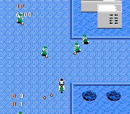 A top-down vehicle combat scene displays a player-controlled green jeep in the center of a blue tiled road, with three enemy green jeeps positioned around the playfield. The HUD shows a score of 00 in the upper left. A gray building structure appears in the upper right corner. Two dark circular objects, likely mines or obstacles, are visible in the lower right section. The sprite-based graphics use a limited palette characteristic of NES-era shooters, with pixelated vehicles and terrain rendered in bright blue tones.