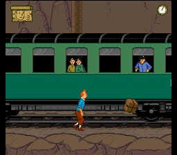 A character sprite stands on a train platform rendered in brown and tan earth tones. A green freight train with multiple cargo cars spans the upper portion of the screen, with two characters visible in separate windows of the train. The character on the ground wears blue clothing and faces the train. A small icon in the upper-left corner shows an inventory or status image. The 16-bit SNES graphics display typical sprite-based platformer styling with parallax background elements showing the ground texture beneath the train platform.