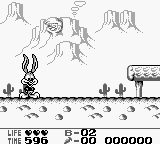 A Game Boy screenshot shows a desert level with a small character sprite on the left side of the screen standing on sandy ground with cacti. The background displays rock formations and a distant landscape with parallax scrolling. The top of the screen displays the HUD with "LIFE ★★★★" on the left, a timer showing "02" minutes and "86" seconds in the center, and "000000" for the score on the right. The sprite is rendered in monochrome pixel art typical of Game Boy graphics.