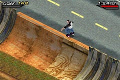 A skateboarder in blue clothing performs a trick atop a large wooden ramp structure with brown and tan coloring. The ramp has curved edges and sits against a gray asphalt surface with yellow lane markings visible in the background. The top-left corner displays a score counter reading "0.00 x 1.0", while the top-right shows a timer at "1:24 PM". The graphics use a low-resolution sprite style typical of Game Boy Advance hardware, with simplified geometry and a limited color palette.