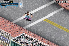 A skateboarder performs a trick in mid-air above an urban street intersection. The isometric viewpoint shows concrete pavement with yellow and white lane markings, red brick stadium seating in the background, and a visible score display in the upper left corner showing a numeric value. The sprite-based graphics use a limited color palette typical of Game Boy Advance hardware, with pixelated character and environmental details rendered in the system's characteristic low resolution.