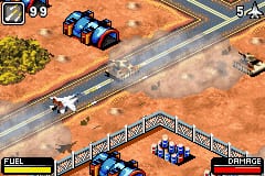 An overhead isometric view of a military base layout with brown terrain and gray paved roads. A blue armed helicopter occupies the upper-left area near a small structure, while white and blue military vehicles are positioned on roads and landing pads throughout the scene. The HUD displays 99 ammunition in the upper-left corner, with a fuel bar and bridge indicator visible at the bottom. The sprite-based graphics use a limited color palette typical of Game Boy Advance hardware, with distinct pixelation and flat shading on all game elements.