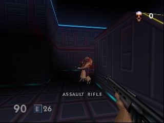 A first-person perspective shows a player character holding an assault rifle in a futuristic corridor with dark metallic walls and blue neon trim. The HUD displays "ASSAULT RIFLE" as the equipped weapon, with ammunition count "96" and health "90" shown in the lower left. A brown humanoid enemy figure stands ahead in the hallway. The environment uses dark colors with geometric architecture typical of N64-era 3D rendering.