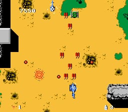 A top-down scrolling shooter displays a yellow desert landscape dotted with brown and green enemies scattered across the screen. A blue and white helicopter sprite appears in the center-lower portion of the game field. Multiple brown circular enemy sprites with radiating patterns are positioned throughout, along with smaller green square structures. A black vertical strip forms the left screen edge. Small red diamond-shaped projectiles or pickups are visible in various locations. The color palette consists primarily of yellow background with brown, green, and blue sprite elements.