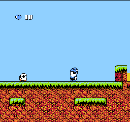 A side-scrolling platformer level displays a light blue sky above a green grass line. Two small white characters with round heads stand on red brick terrain scattered with green grass tufts. A heart icon and the number 10 appear in the upper left corner, indicating health or lives. On the right side, a colorful enemy sprite with red and yellow coloring stands near the level's edge. The scene uses NES-era pixel art with a limited color palette and 8-bit sprite resolution.