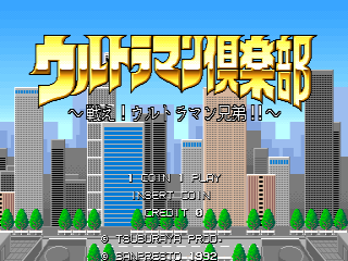 The title screen displays Japanese text in large yellow pixelated letters across the top, with a cityscape of tall buildings in the background under a blue sky. Green bushes and trees line the bottom of the screen. Below the title, menu options and credits text are visible in smaller white Japanese characters, arranged in multiple rows. The overall scene uses a flat, side-scrolling arcade aesthetic with bright primary colors and clear sprite-based graphics.