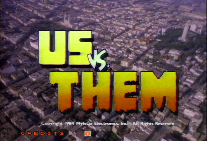 The title screen displays "US vs. THEM" in large yellow and orange gradient text centered over an aerial cityscape photograph. A small "vs." separator appears between the two words in white. Below the title, copyright information reads "Copyright 1984 Mylstar Electronics, Inc. All Rights Reserved" in small white text. The word "CREDITS" appears in red text in the lower left corner, with a small orange square indicator to its right. The background shows an overhead view of an urban area with buildings and streets in muted purple and brown tones.