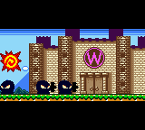 A 2D side-scrolling level displays Wario as a small sprite on the left side of a grassy platform. Brown brick structures and a tall castle-like building with a purple 'W' emblem form the background. The sky is light blue at the top. A yellow sun appears in the upper left corner, and black enemy sprites are visible on the ground. The pixel art uses a limited Game Boy color palette of greens, browns, and blues typical of early handheld game graphics.