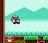 Wario, rendered as a small pixelated sprite with red and yellow coloring, stands on a green platform in the left-center area of a side-scrolling level. Behind him rises a teal-colored mountain landscape against a light cyan sky. The foreground shows a dark brown brick ledge with green block segments, typical of platformer level design. The Game Boy's limited color palette of greens, browns, and pastels is clearly visible throughout the scene.