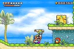 Wario stands on a sandy beach in the foreground, facing right toward a green platform suspended above water. A palm tree rises on the left side, and the ocean and sky with white clouds fill the background. The HUD displays Wario's health as four filled squares in the top-left corner, a coin counter showing 000360 in the top-right, and a red spherical enemy sits on the sand to the right. The scene uses bright, solid colors typical of Game Boy Advance sprite graphics with a tropical daytime setting.