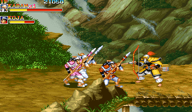 Three sprite-based warriors in colorful armor engage in melee combat on a tan battlefield with rocky outcrops and green grass visible at screen edges. The leftmost character wields a sword while standing, the center character crouches with a weapon, and the rightmost character strikes with extended reach. A brown wooden log or branch extends across the lower portion of the screen. The top-left corner displays two player health bars with character portraits and a score readout showing 21050. The scene uses a side-scrolling perspective with a parallax background of layered terrain and waterways.