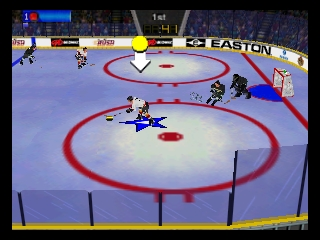 An overhead view of an ice hockey rink with red circles marking the playing surface and blue boards at the edges. Two players in blue jerseys position themselves on the ice, with one player near center ice. A yellow puck sits in the middle area. The UI displays player health bars and team names at the top, including "EASTON" branding visible along the boards. The graphics show N64-era 3D polygon rendering with flat textures and low polygon counts typical of late-1990s hardware.