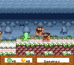 A colorful sprite-based game scene shows a green character with a round body in the center-left of the screen, positioned on a grass platform with white flowers. Brown wooden platforms with soil textures form the ground level below. The background displays a blue sky with repeated jellyfish-like sprites arranged in rows at the top. A UI panel at the bottom contains two resource indicators on the left (a green circle showing 56 and an orange circle showing 0), with the word "Sandros" displayed in the center-right. The overall aesthetic uses bright colors typical of SNES-era pixel graphics with clear parallax layering between background and foreground elements.