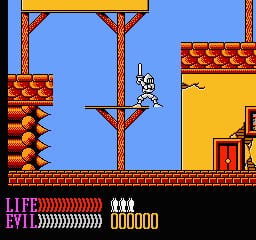 A blue-sky platformer level features the protagonist Kuros suspended from a rope in the center, positioned between wooden scaffolding structures. Red and orange brick platforms with decorative patterns line the left and right sides. The bottom of the screen displays a black HUD bar with white text showing "LIFE" and "EVIL" meters on the left in red and orange, and a gold coin counter "000000" on the right. A small red fortress structure appears in the lower right corner against a yellow background.
