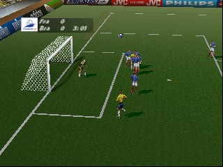 A soccer match in progress on a green pitch viewed from an overhead perspective. A player in blue and white striped jersey stands near the goal area on the left, while a goalkeeper in yellow is positioned in the net. Another blue-and-white player and additional team members are scattered across the field. White field markings, the goal line, and penalty box are visible. Sponsor boards with logos line the top edge of the screen, including JVC and Philips branding. The 3D polygonal graphics and field layout are characteristic of late-1990s sports game rendering.