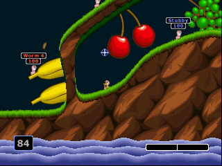 A yellow worm character stands on a green vine branch in the upper left, facing three red apples hanging above. Brown rocky terrain dominates the right side, with water visible at the bottom. The HUD displays a score of 84 in the lower left corner and a health bar at the bottom center. Text labels show weapon or item names in the upper corners. The art style uses bright, solid colors with pixelated sprite graphics typical of late-1990s console gaming.