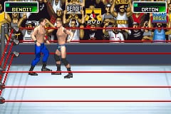 A wrestling match is in progress on a blue and white squared ring. Two wrestlers grapple in the center, with one in blue trunks and the other in red. Pixelated crowd sprites fill the background stands in shades of red and yellow. Health bars and player names appear at the top of the screen in yellow and green text boxes labeled with wrestler identities. The art style features low-resolution 2D sprites typical of Game Boy Advance graphics.