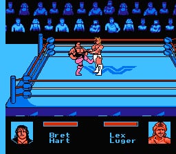 Two red-and-flesh-colored wrestler sprites face off in the center of a blue wrestling ring, viewed from above. A row of blue-pixelated spectator sprites lines the top edge of the ring. The lower third displays a black UI panel with two wrestler portraits on either side—the left showing a character labeled "Bret" with a health bar above, the right showing "The Loser" with a corresponding health bar. The arena background uses a light blue sky with darker blue structural elements framing the wrestling ring.