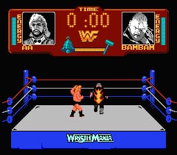 A wrestling ring occupies the center of the screen with two pixelated wrestlers facing each other on a white mat bordered by blue ropes and blue corner posts. The top of the screen displays a scoreboard with two wrestler portraits in boxes on either side—one labeled with a skull and one labeled "KAMDAN"—separated by a red "W" logo and digital time display showing "0:00" in the middle. The bottom shows a blue banner reading "WRESTLEMANIA" in white text. The background is dark with red trim framing the match view.