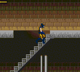 A pixelated character sprite in blue and yellow stands on a diagonal brick staircase in the center of the frame. The staircase connects two brown brick platforms against a dark background. A brown brick wall with lighter horizontal bands fills the upper portion. Two vertical cylindrical pipes in gray and olive green rise from the lower edge. The overall color palette is muted earth tones with limited sprite detail typical of Game Gear hardware.