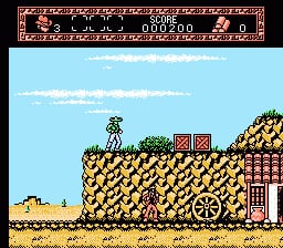 A side-scrolling platformer view shows a character standing on tan-colored rocky terrain in the center-left of the frame. The landscape features yellow and brown pixelated rocks and earth formations, with a red-brick structure visible on the right side. The bright blue sky fills the upper portion with white clouds. A small helicopter appears in the upper-left corner. The NES-era sprite graphics display limited color palette and low resolution typical of early 1990s cartridge games.