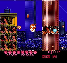 A large pixelated giant head with an open mouth dominates the center of the screen against a blue night sky with tall buildings and windows on either side. Pink and red enemy sprites line the bottom of the screen in a horizontal row. The left side shows an orange building with decorative elements, while the right features a tall brick-textured tower. A score display appears at the bottom in red text, and the overall color palette uses bright reds, oranges, yellows, and blues against a black ground area.