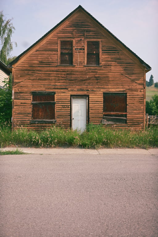 A weathered timber building with boarded windows, a white door and tall grass along the street.