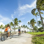 Individuals of all ages cycling through Lummus Park along the Miami Beach Boardwalk.