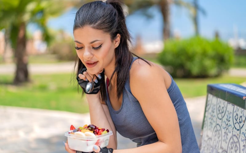 A young, fit woman is holding a bowl of fruit outdoors, preparing to enjoy a healthy snack after her workout