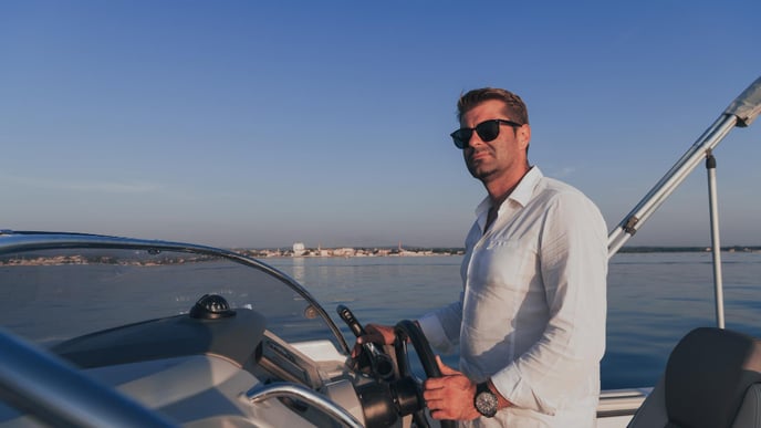 A focused senior executive, dressed in casual attire and sunglasses, relishes his vacation as he navigates a luxury boat during sunset