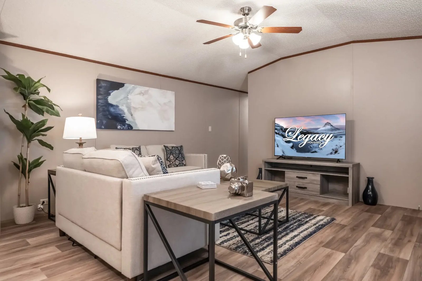 Living room in The Lisa by Legacy Housing with white seating, a television area, ceiling fan, and wood-look flooring
