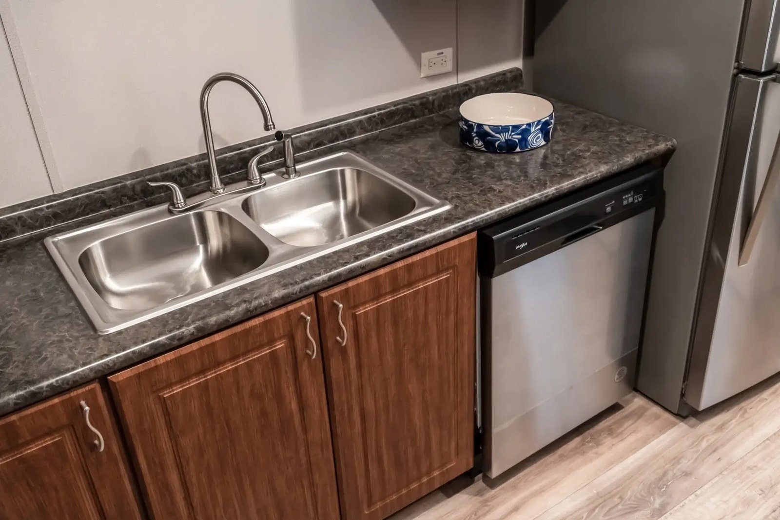Kitchen sink area in The Lisa with a stainless double-basin sink, dishwasher, dark countertop, and refrigerator