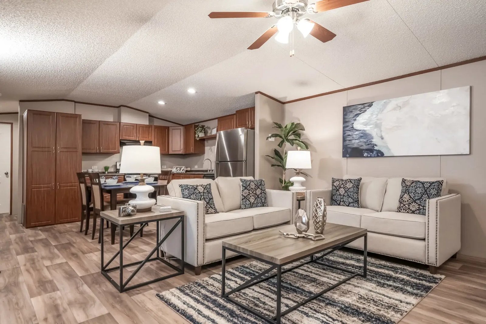 Open living room, dining area, and kitchen in The Lisa by Legacy Housing with wood-look flooring and warm wood cabinetry