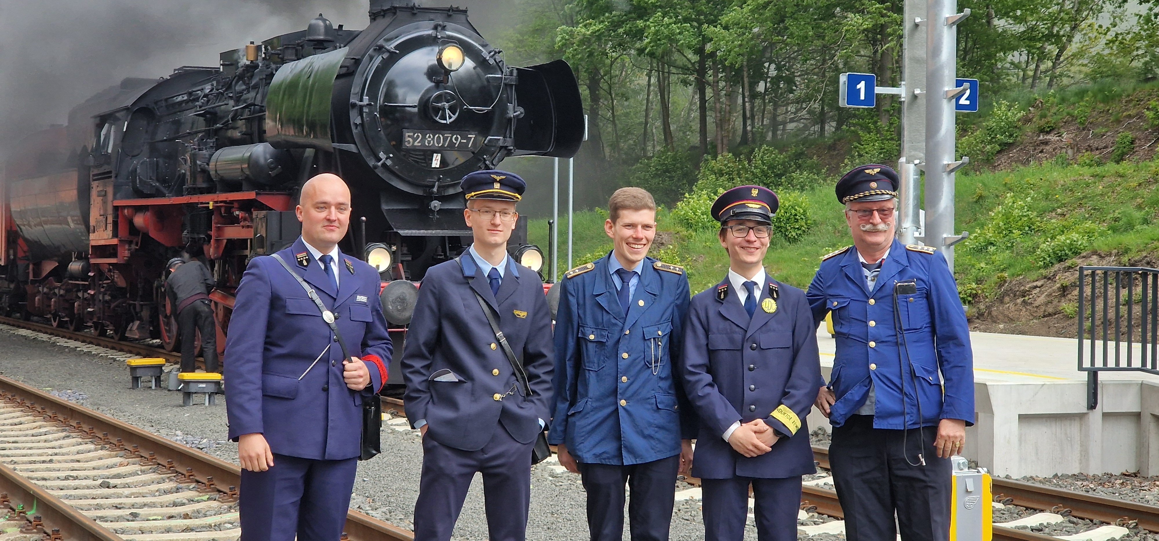 Pięciu kolejarzy w mundurach stoi w rzędzie, w tle parowóz BR52
