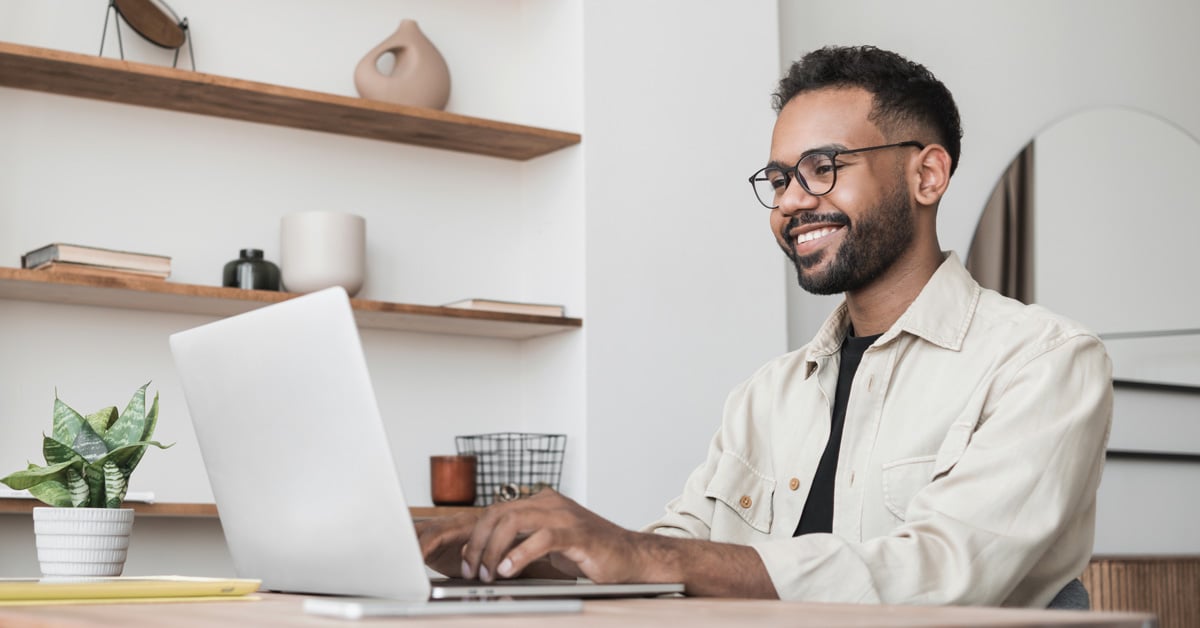 A man wearing glasses and a light shirt smiles while working on a laptop at a desk. Shelves with books and decorative items are in the background, along with a small potted plant beside him as he prepares to request info online.