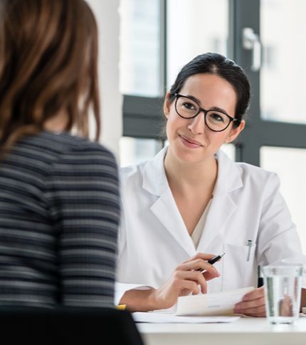 A female Physician Office Specialist in a white coat and glasses smiles while holding a pen and notepad, talking to a patient across the desk in a bright office setting.