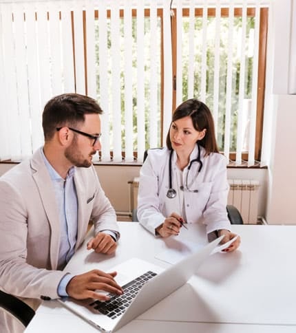 A doctor in a white coat and stethoscope talks to a man in a suit at a desk. The man uses a laptop while they both review a document. They are in a bright office with large windows and vertical blinds.