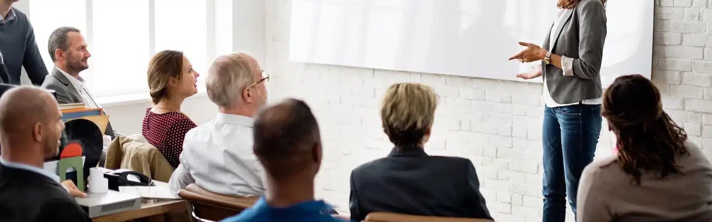 A person is giving a presentation to a group of attentive people seated in a room with a large whiteboard.