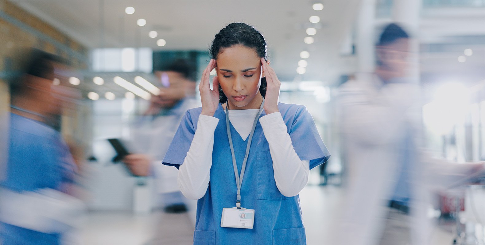 A healthcare worker in scrubs stands still with her hands on her temples, looking stressed, as blurred figures of other medical staff move quickly around her in a busy hospital corridor.