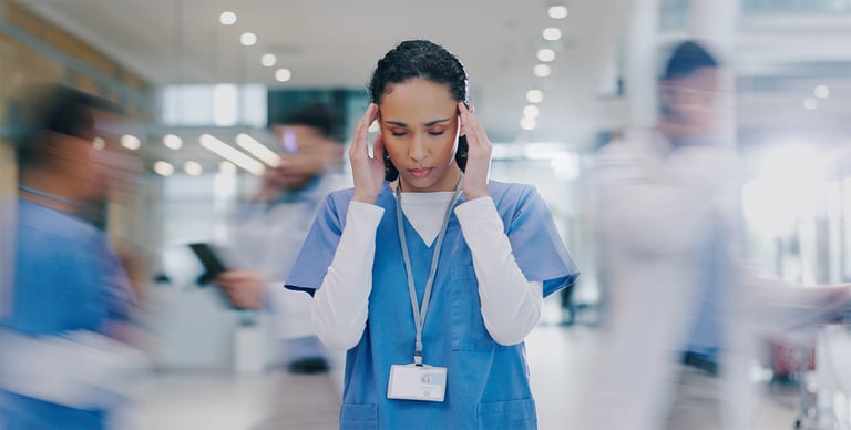A healthcare worker in scrubs stands still with her hands on her temples, looking stressed, as blurred figures of other medical staff move quickly around her in a busy hospital corridor.