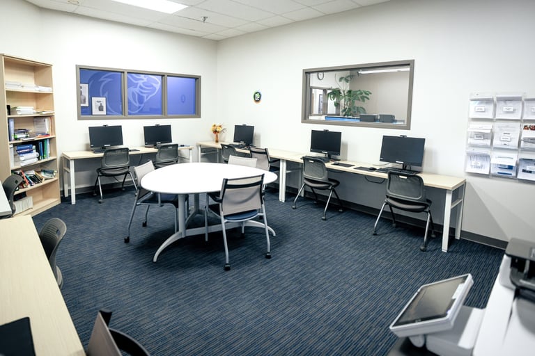A clean, modern computer lab in Henrietta, NY features several desktop computers on desks, a round table with chairs in the center, bookshelves lining one wall, expansive windows letting in natural light, and a blue carpeted floor.
