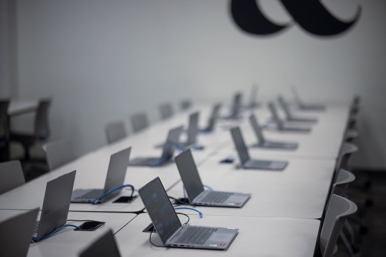 A row of open laptops on a long white table in an empty, modern classroom or office with gray chairs and a partial black wall design visible.