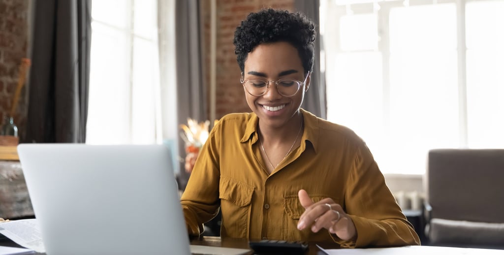 A person wearing glasses and a mustard-colored shirt sits at a desk, smiling while working on a laptop—perhaps inspired to enter the field during this accountant shortage. They use a calculator, with papers and a pen on the desk in a well-lit room.