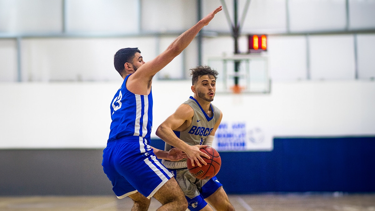 Two basketball players in a brightly lit gym, one in a blue jersey playing defense against another in a grey jersey holding the ball.