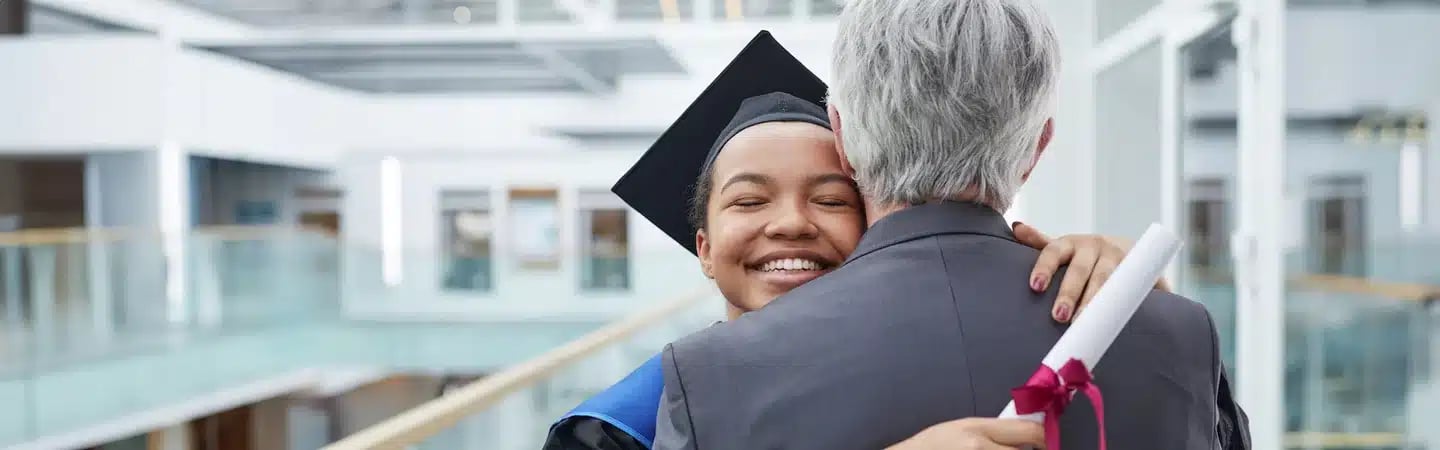 Graduate in cap and gown hugging an older person while holding a diploma with a red ribbon in a modern building.